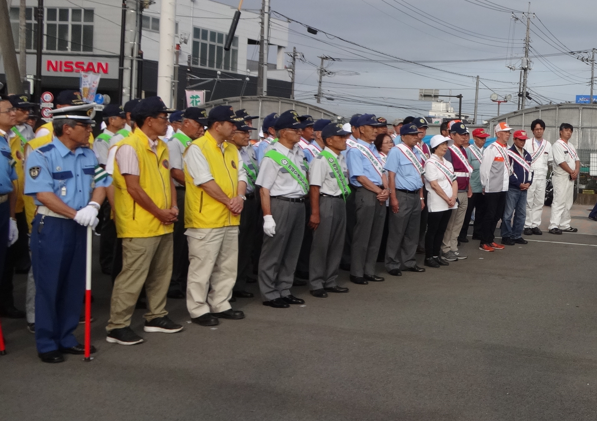 警察官や地域の防犯ボランティアなどが大勢の人々が、制服や黄色のベスト、腕章やタスキを着用して整列している写真