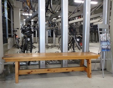 駐輪場のコンクリートの床の上に、明るい茶色の木製ベンチが正面に置かれ、その奥に複数の自転車が二段式の駐輪ラックに整然と格納されている様子を写した写真