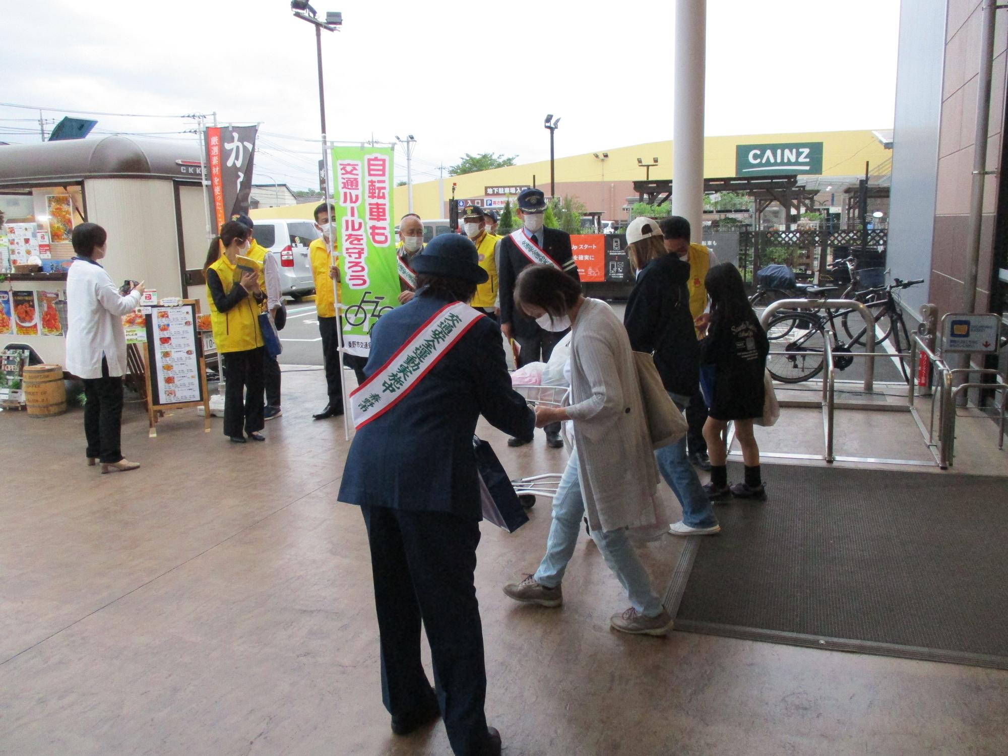 黄色のベストを着用したボランティアの人々が交通安全を呼びかけるのぼり旗を持ち、出入口の左右に立つ交通安全のタスキをかけた制服姿の2人の警察官が通行人にチラシを配布している活動の様子の写真