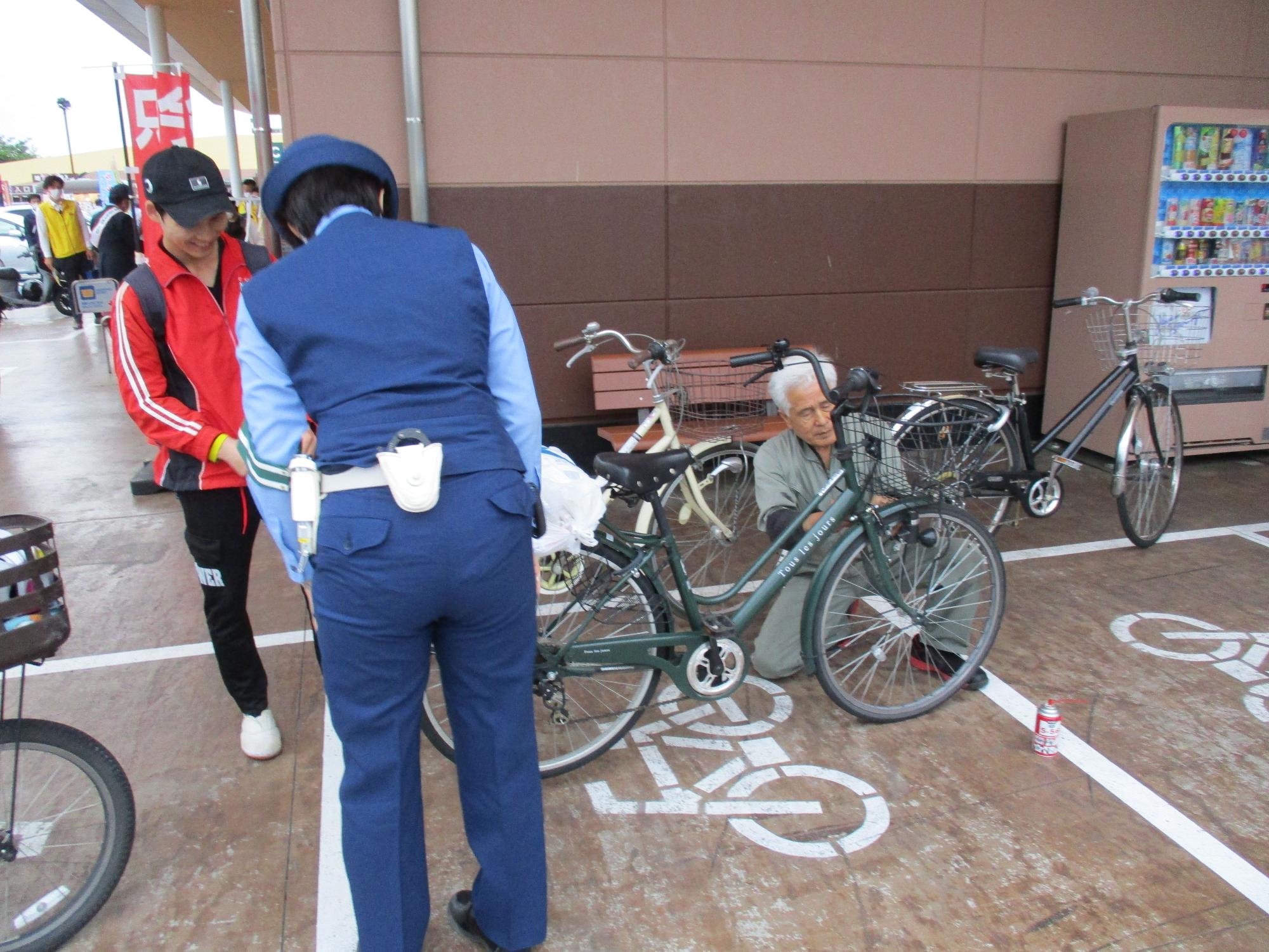 駐輪場で、緑色の自転車の前輪を男性が見ており、その横では赤と黒のスポーツウエアを着た笑顔の女性と制服姿の女性警察官が見守っている交通安全キャンペーンの写真