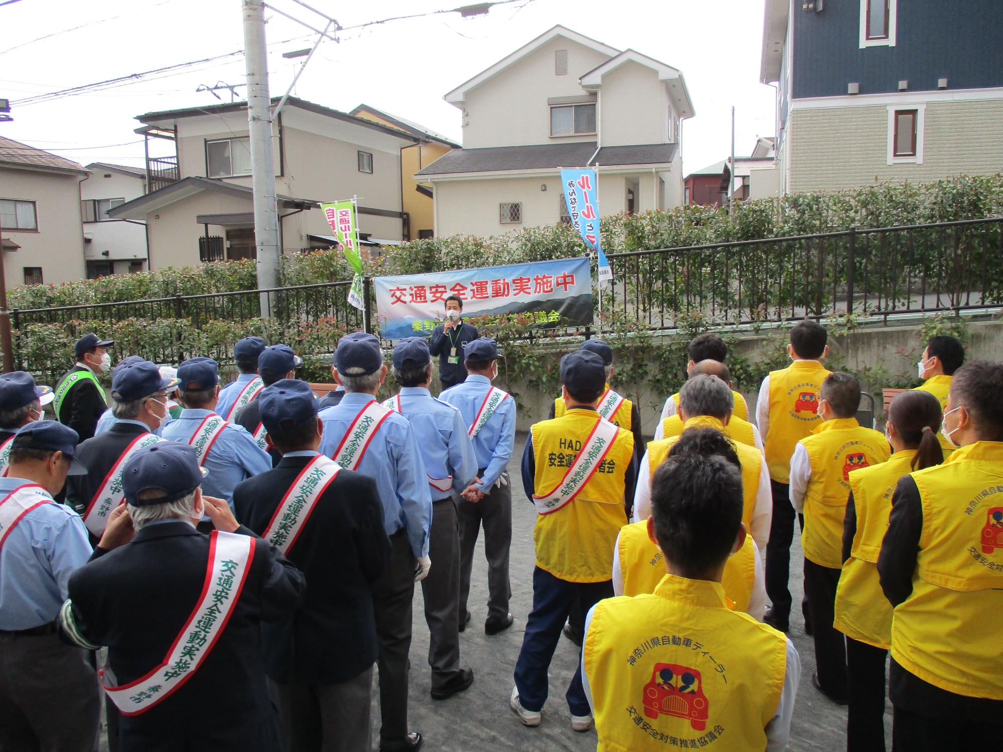 黄色のベストを着用した地域ボランティアと、たすきに青色のシャツを着た参加者たちが整列し、前方に立ってマイクを持って発言している一人の男性の話を聞いている様子を捉えた交通安全運動の出発式の様子の写真