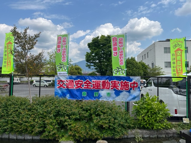 駐車場の境目に間隔をあけて設置されたのぼり旗と、柵に「交通安全運動実施中 秦野市」と書かれた横断幕が掲げられている写真