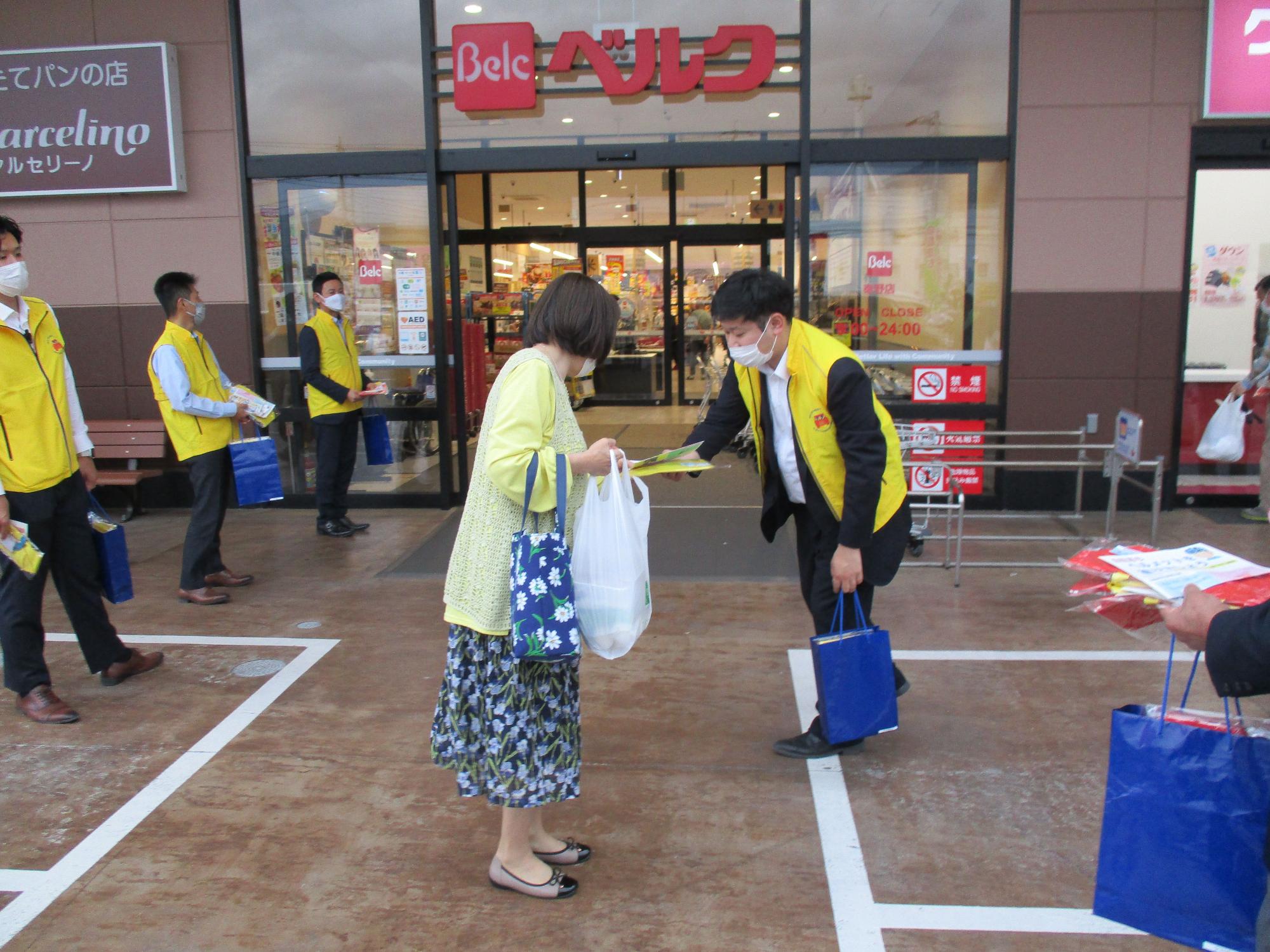 スーパーマーケット「ベルク」の入り口前で、黄色のベストを着た複数の男性が並び、右側の男性が花柄のスカートを着た買い物客の女性に青い袋に入ったチラシや啓発品を配っている様子を捉えた写真