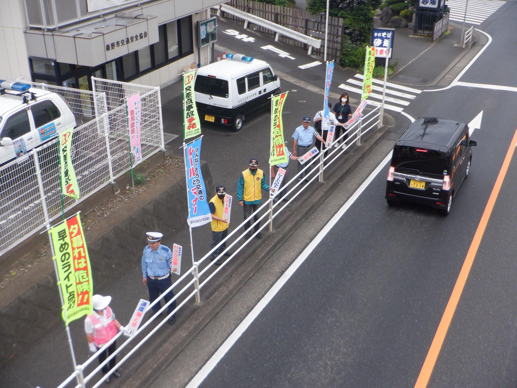 緑と青色ののぼり旗が交互に立っている道路沿いの歩道に2人ずつ並び、交通安全の小さな旗を持った制服姿の警察官やボランティアの人々が、行き交う車や通行人に呼びかけをしている交通安全啓発活動の様子の写真
