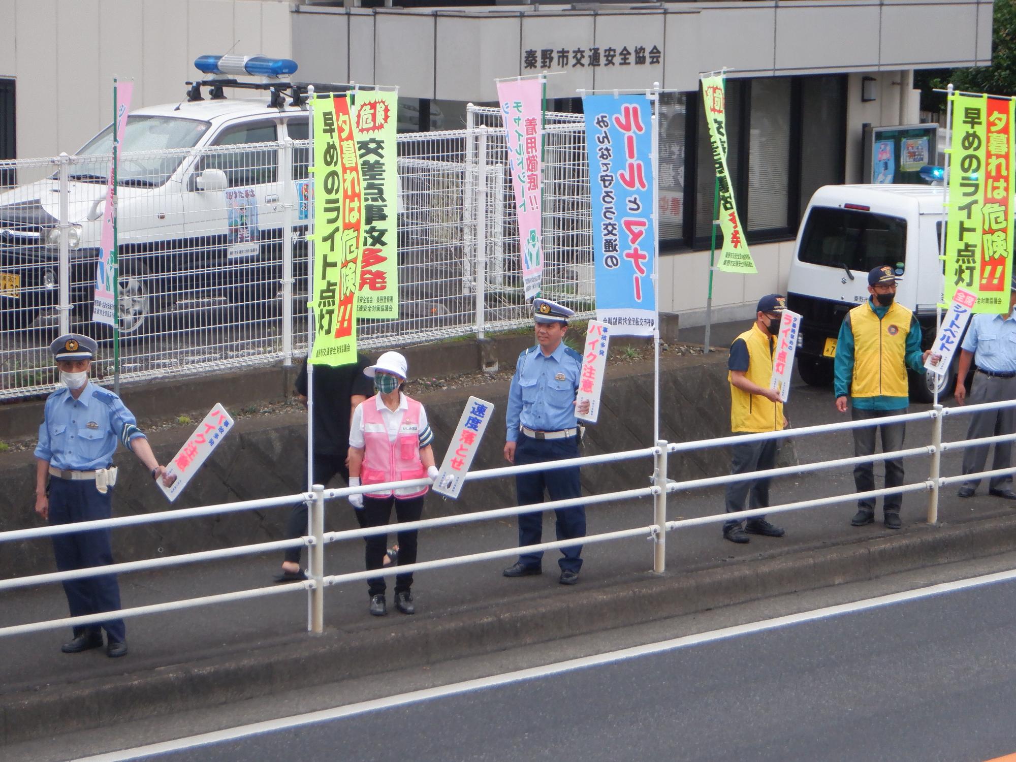屋根の上に青色のパトランプがついた白と黒の車が停車してる「秦野市交通安全協会」と書かれた建物前の歩道脇で、制服姿の複数の警察官やボランティアの人々が、交通安全を呼びかける小さなのぼり旗を持って立ち、行き交う車にアピールをしている様子を捉えた写真
