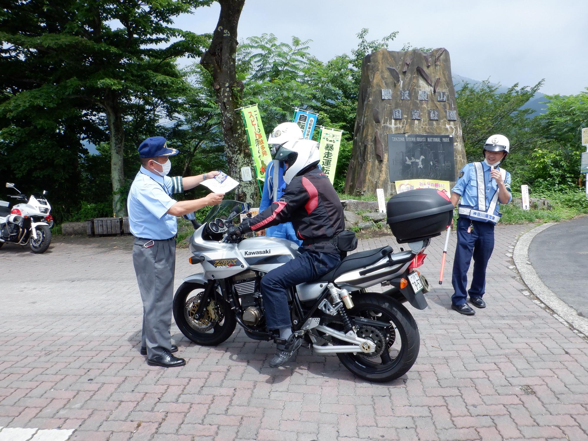頭にヘルメットを被った制服姿の警察官が銀色のバイクに乗った男性を停めて、左腕に緑と白のストライプの腕章をつけた男性が、交通安全についてのチラシと啓発品を持って説明をしている様子を、道路側に立っている警察官が見ている屋外の交通安全啓発活動の様子の写真