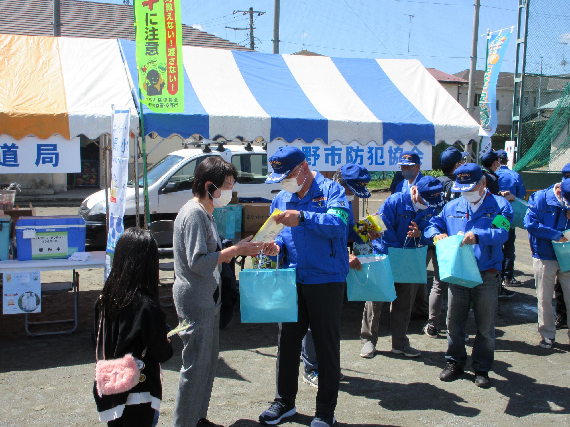「秦野市防犯協会」と書かれた青と白のストライプのテントの前で、マスクを着用し青いキャップとジャンパーを着た防犯指導員と思われる男性たちが、マスクを着用した女性に対して、青い紙袋に入った啓発品を説明しながら手渡している様子の写真