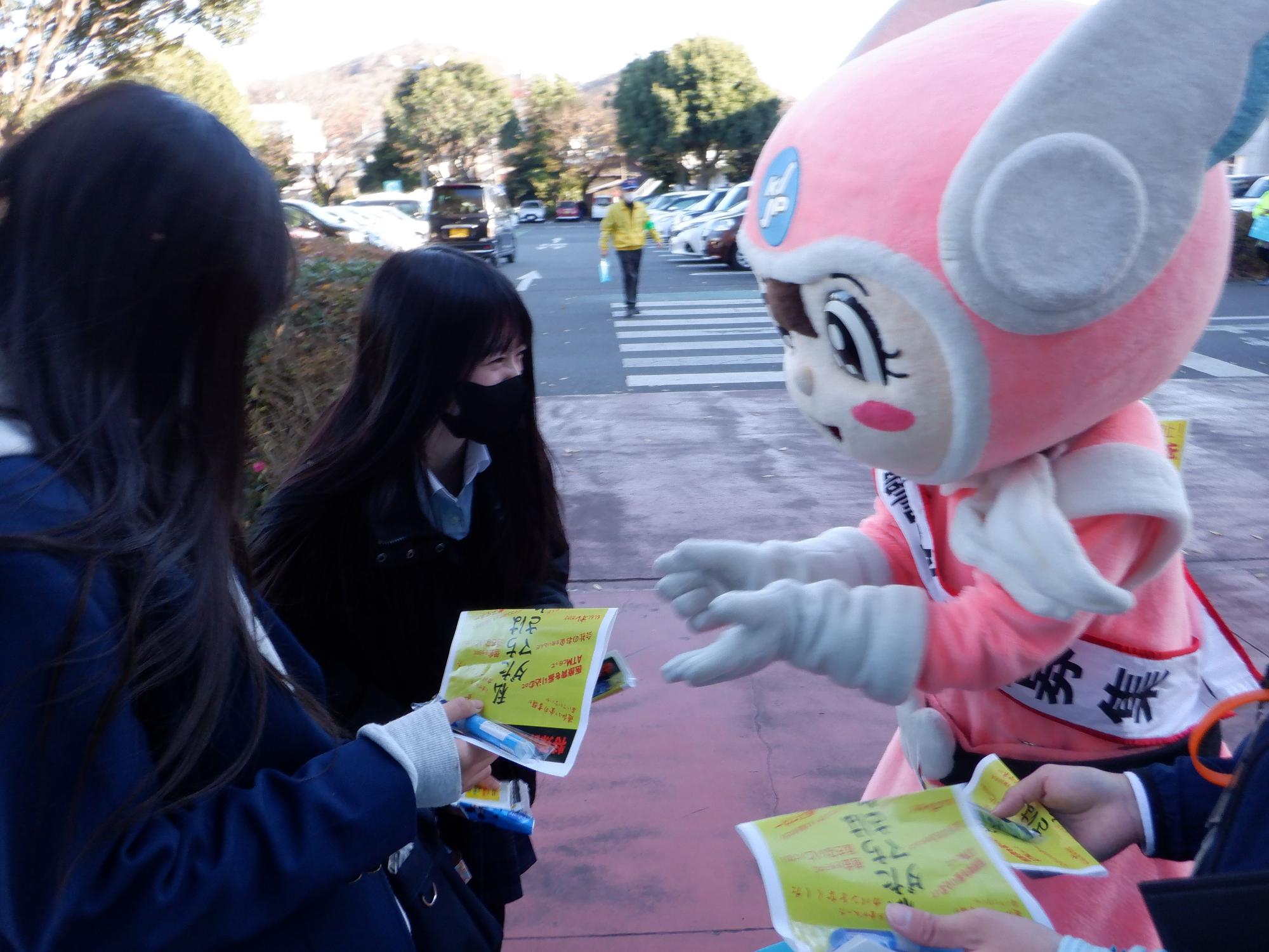 頭上に羽のような飾りがついたピンク色のキャラクターが、マスクを着用した2人の女性生徒に啓発品とチラシを配布して啓発活動を行っている様子の写真