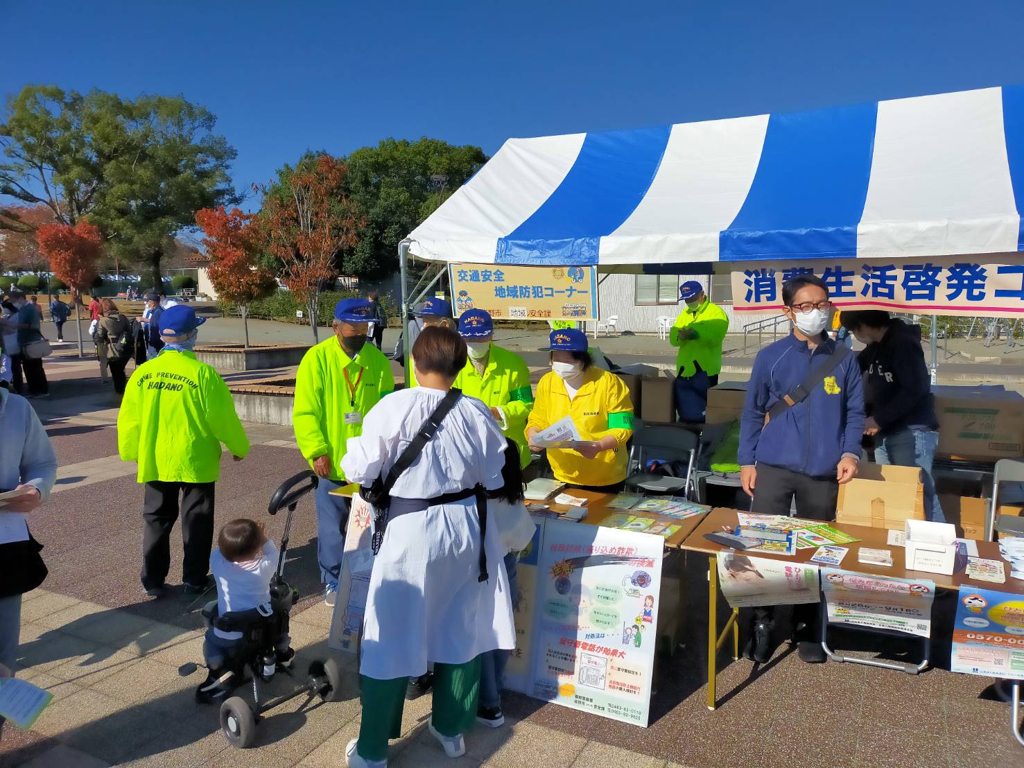天気の良い広場で、「交通安全地域防犯コーナー」と書かれた青と白のストライプのテントの前で、黄緑や黄色いジャンパーと青い帽子を着用したスタッフが、親子連れの来場者に向けて防犯に関するチラシを配っている様子の写真