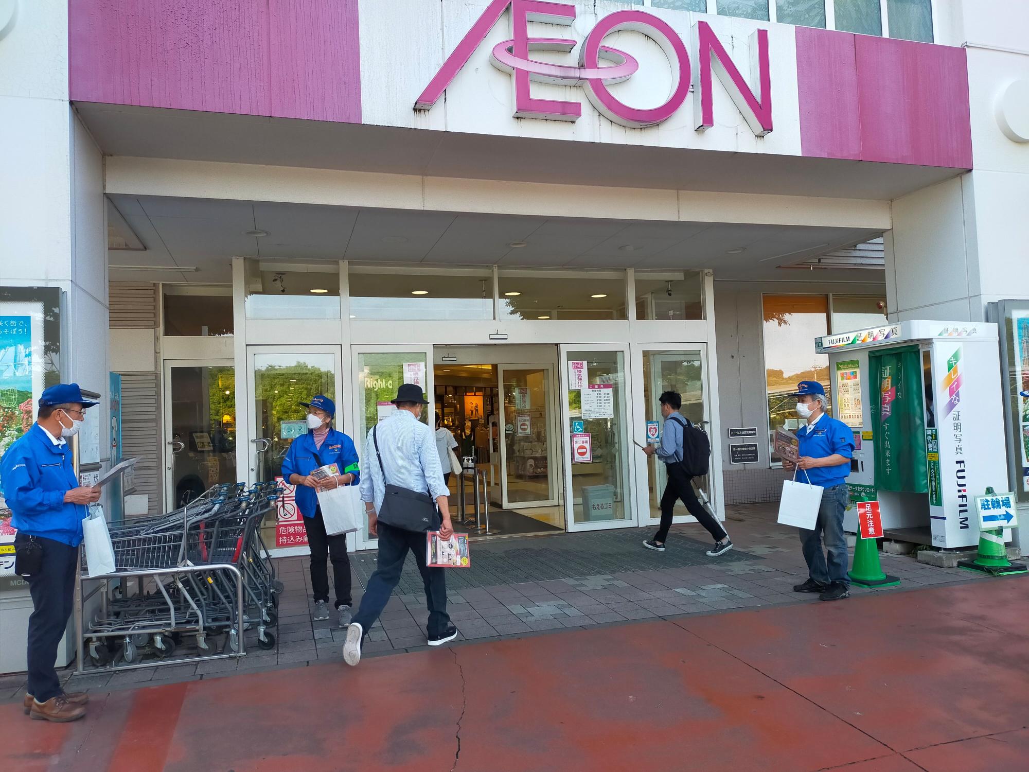 AEONの店舗入り口前で、青いジャンパーにキャップを被ってマスクをした3人のスタッフが、買い物客にチラシなどを手渡し防犯啓発活動を行っている様子の写真