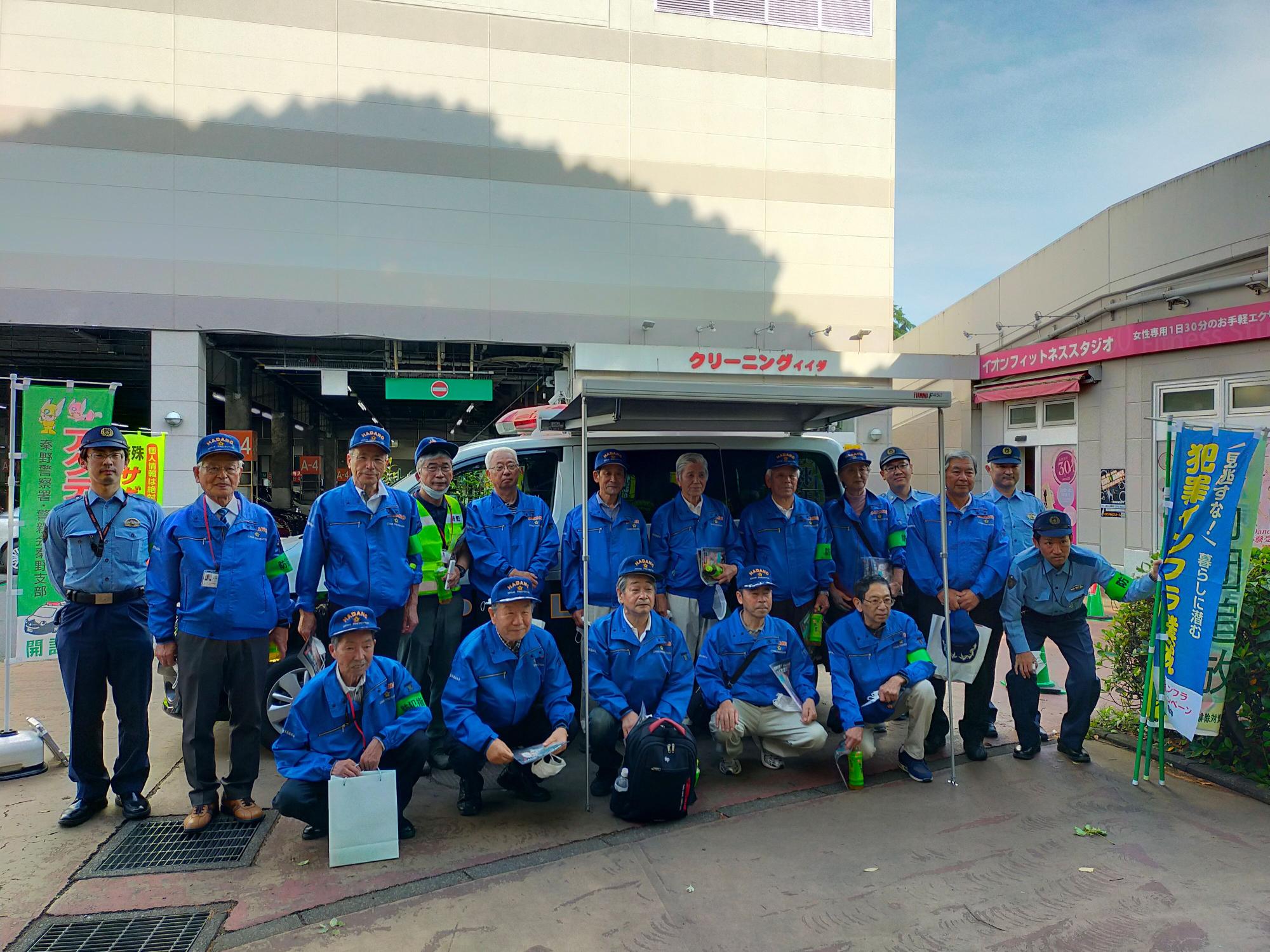 屋根付きの駐車場前の屋外で、制服姿の4人の警察官と青いジャンパーに帽子を着用した複数の男性スタッフがカメラに向かって2列に並んでいる集合写真