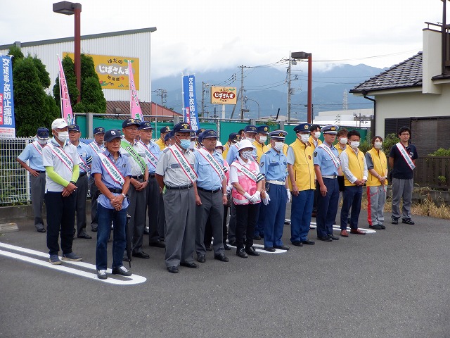 交通安全運動ののぼり旗が設置された屋外の駐車場の一角で、キャップや帽子を被ったタスキを着用した警察官や多くの人々が並んで立っている様子の写真