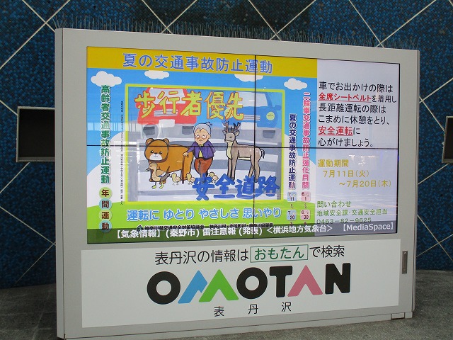 大型デジタルサイネージの画面に「夏の交通事故防止運動」の文字と、「歩行者優先」「安全道路」と書かれたポスターが表示されており、運動期間や交通安全についての注意を促している写真