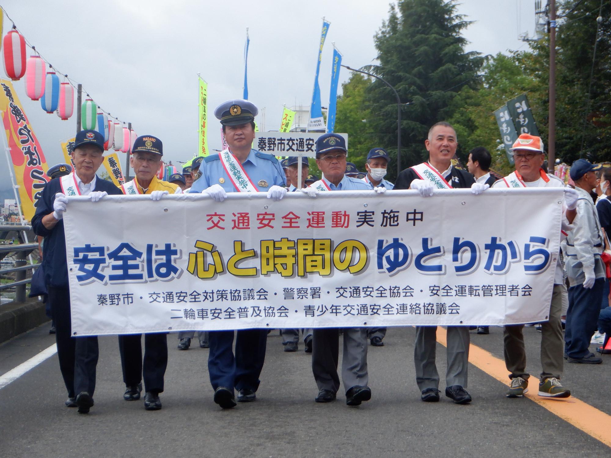 肩にたすきをかけた警察官や関係者たちがスローガンの「安全は心と時間のゆとりから」と書かれた横断幕を持って行進している交通安全運動のパレードの様子の写真
