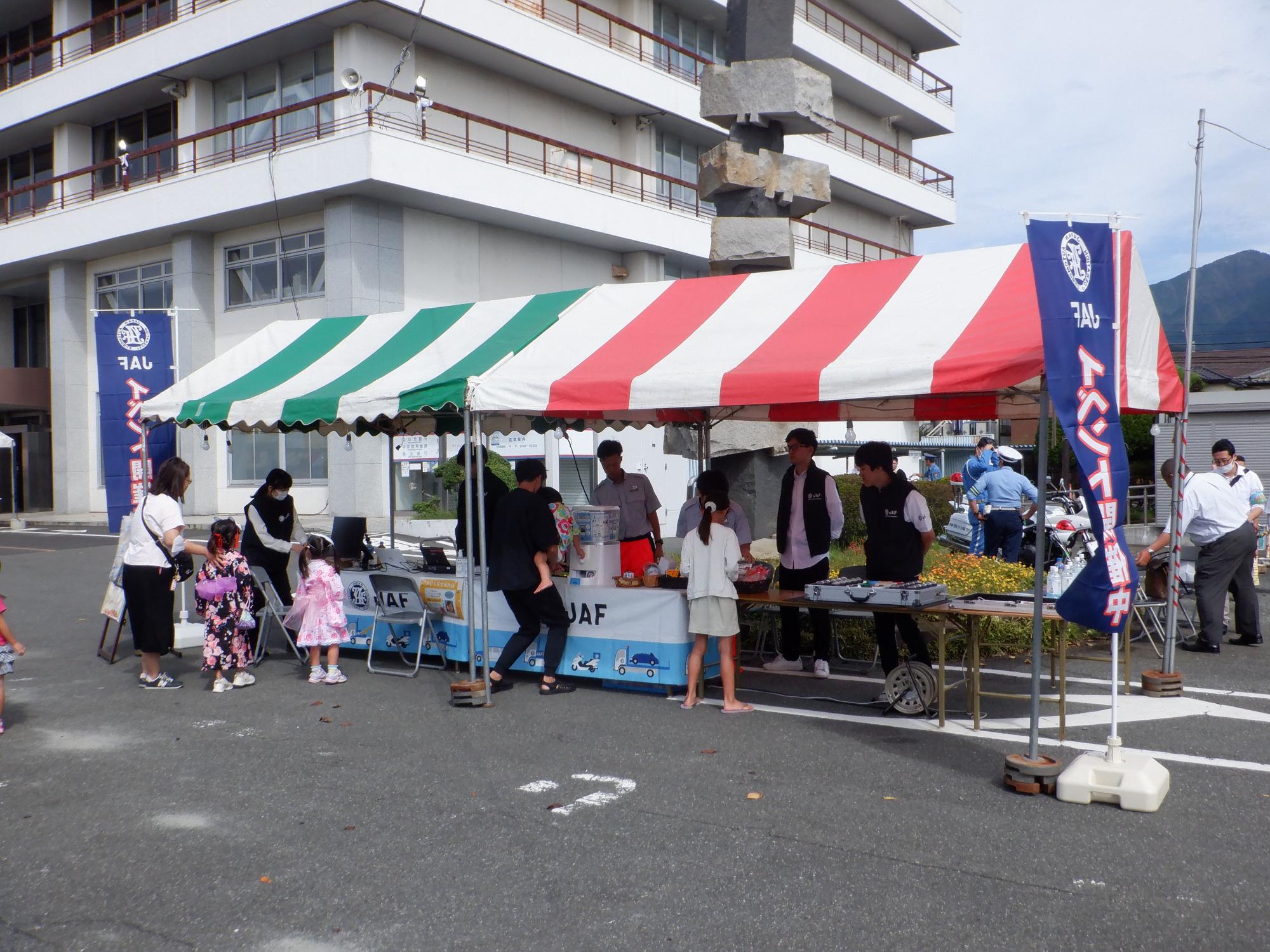 JAFののぼり旗が立つ緑と赤のストライプのテントの下で祭りやイベントの出店が開かれており、男性が抱っこしている小さな子供がガチャガチャを楽しんだり、集まった親子連れの人々が屋台をのぞき込んだりしている屋外イベントの写真
