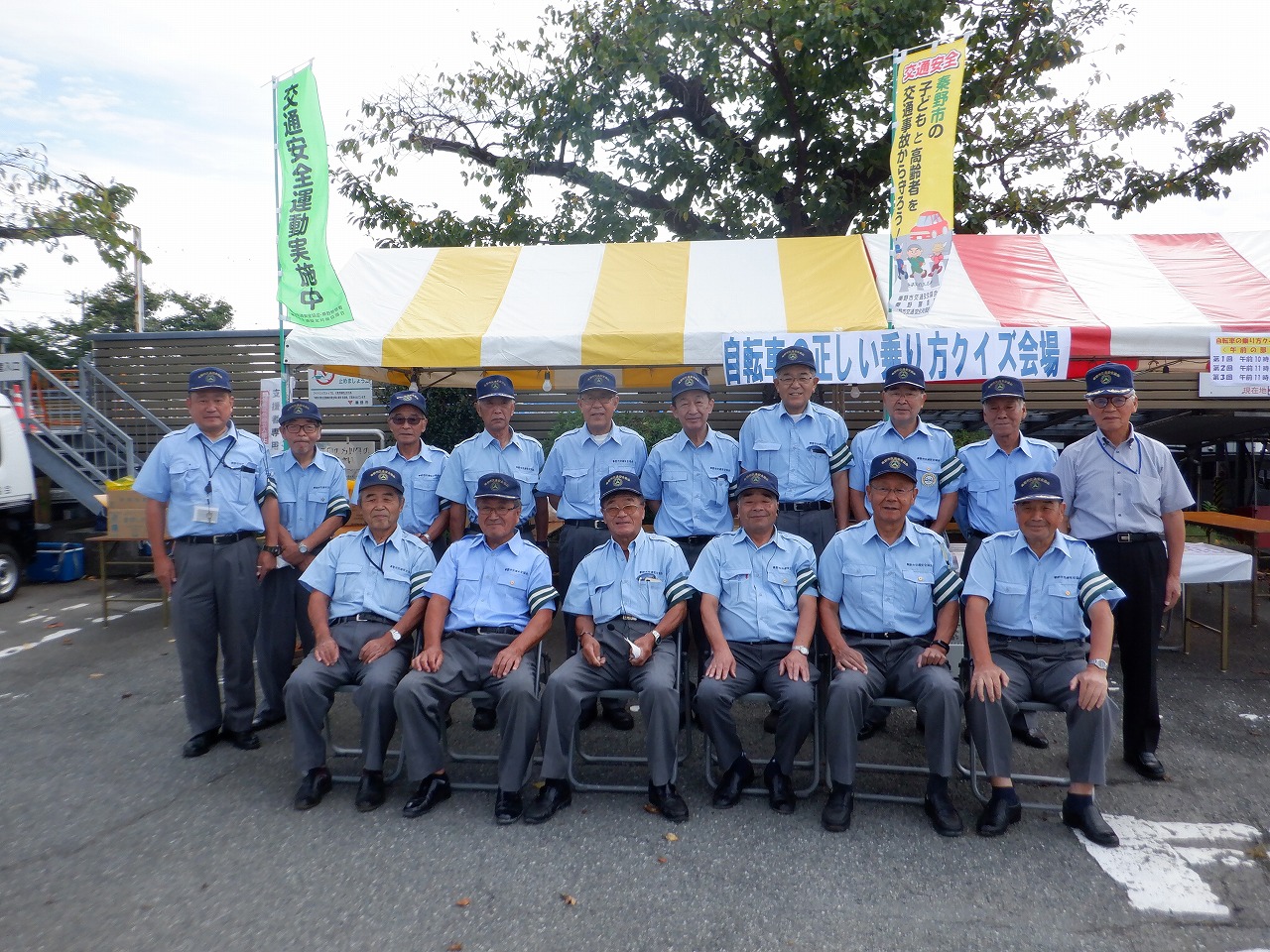 腕に緑と白のストライプ柄の腕章をつけた青い制服を着た大勢の男性が、椅子に座ったり立ったりして2列に並びカメラに笑顔でポーズをとっている集合写真