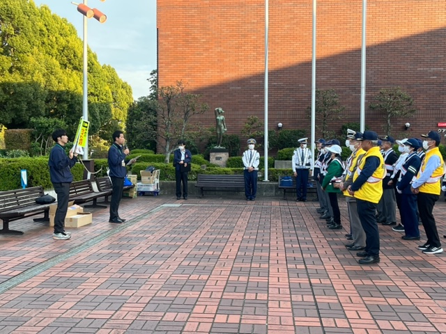 文化会館前の広場に2名の警察官が立ち、手前に大勢の参加者が2列に整列しており、向かい合って立つ男性がマイクを持って話している交通安全運動の出発式の様子の写真