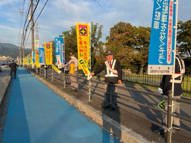 交通安全運動の一環として日が暮れかけた夕方、黄色と青色ののぼり旗が交互に間隔をあけて設置された青い歩道沿いの柵に並んだ複数の男性たちが、走行する車の運転手に向けてそれぞれ小さな旗を持って交通安全を呼びかけている屋外での活動の様子の写真