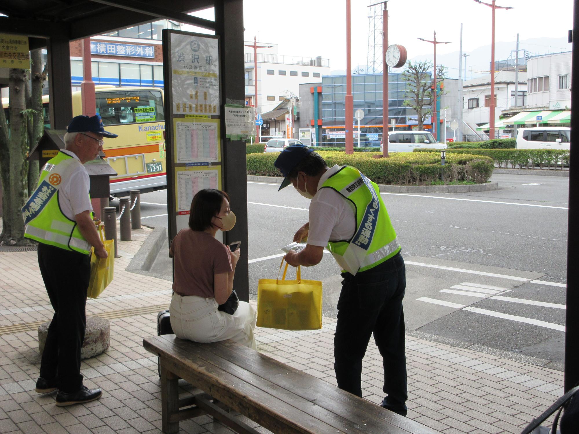 薄い緑色の反射ベストを着用し、青色のキャップを被った二人の男性が、バス停のベンチに腰かけている女性に啓発品を配布している啓発活動の様子の写真
