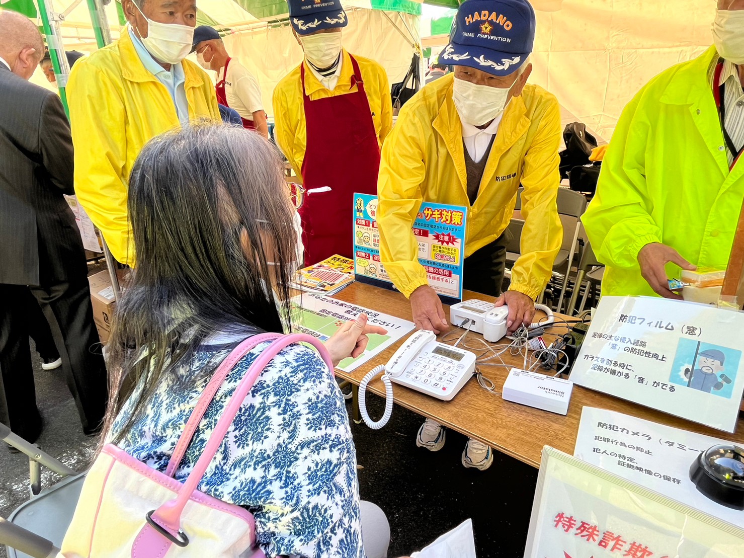 祭りのイベント会場のブースで、花柄のシャツを着た女性がテーブルに置かれた電話機を指差し、黄色のジャンパーを着た男性スタッフから説明を聞いている様子を捉えた写真