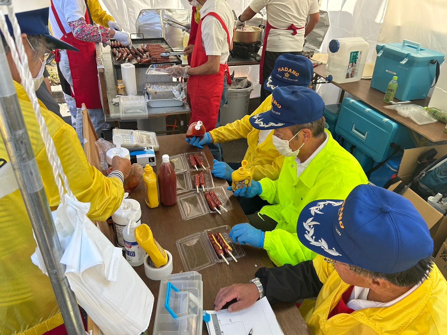祭り会場のテントの下で、黄色いジャンパーと青い帽子をかぶり青色の手袋を両手につけた3人の男性が長テーブルの前に座り、透明のパックに入った焼きたての2本のフランクフルトにマスタードやケチャップをかけている様子の写真