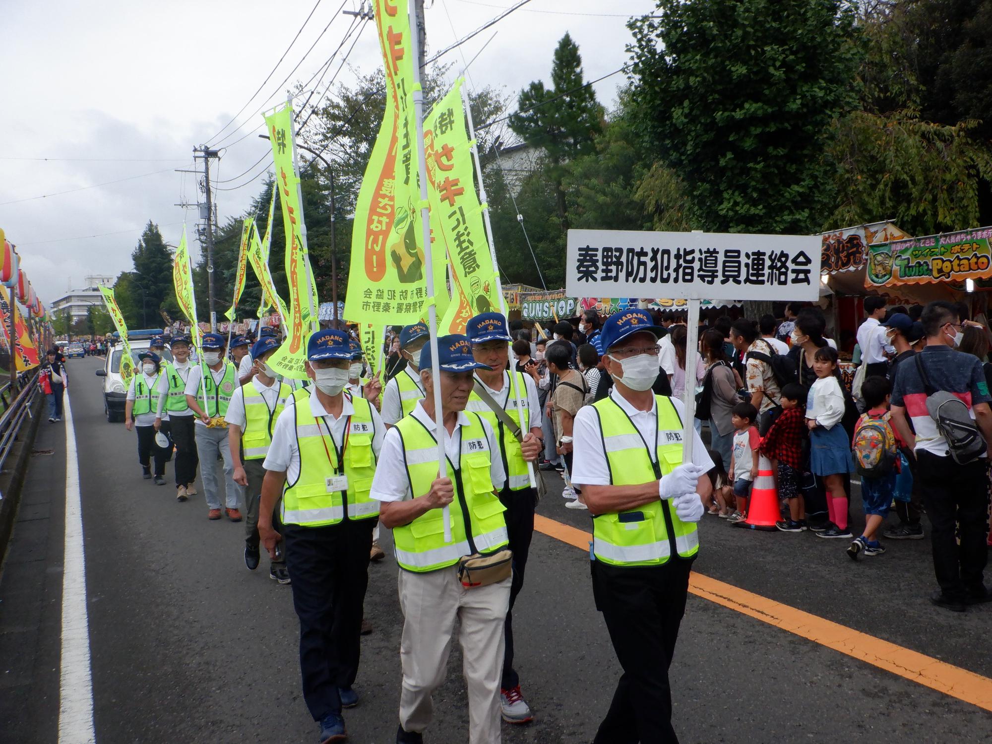 露店が並んでいる歩行者天国になった祭り会場の道路で、反射ベストを着用し青い帽子をかぶった男性たちが、「秦野防犯指導員連絡会」と書かれたプラカードを先頭に、多くの人々で賑わう通りをパレードしている様子の写真