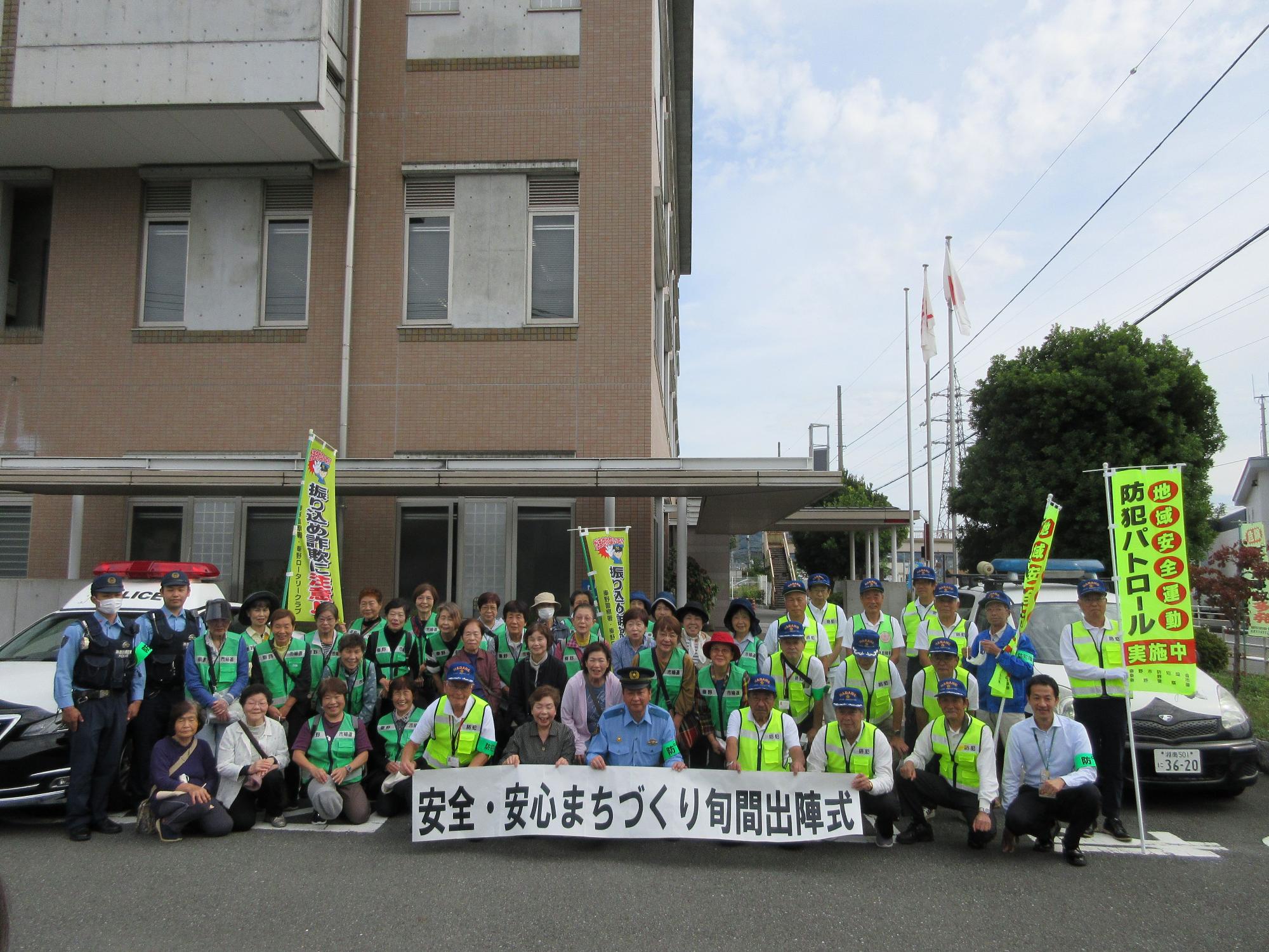 ベージュ色の外壁の建物やパトカーを背景に、2人の警察官と反射ベストを着用した大勢のボランティアが、「安全・安心まちづくり旬間出陣式」と書かれた横断幕を掲げて集まっている集合写真