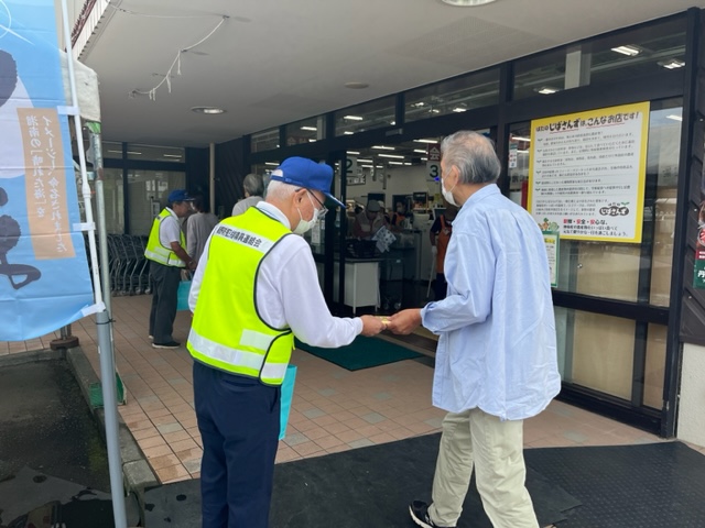 商業施設の建物の入り口前で、青いキャップを被り反射ベストを着た男性が買い物に来た水色のシャツを着た男性客にチラシか何かを渡して防犯啓発活動を行っている様子を捉えた写真