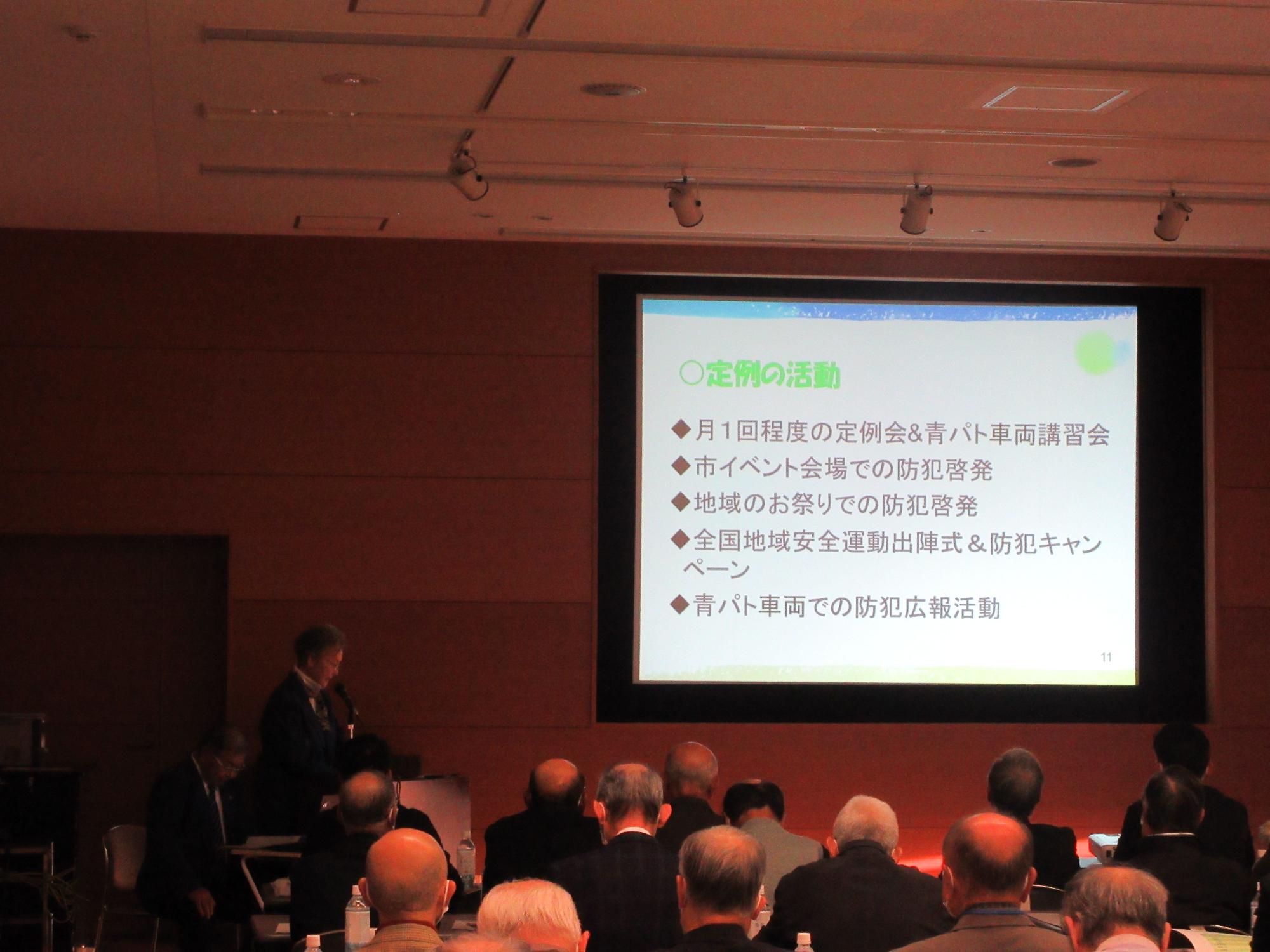 室内の照明が赤く照らされ、前方に資料が映し出されたスクリーンが設置された会議室で、スーツ姿の男性講師が壇上で話す様子を多くの参加者が聞いている研修会の様子の写真