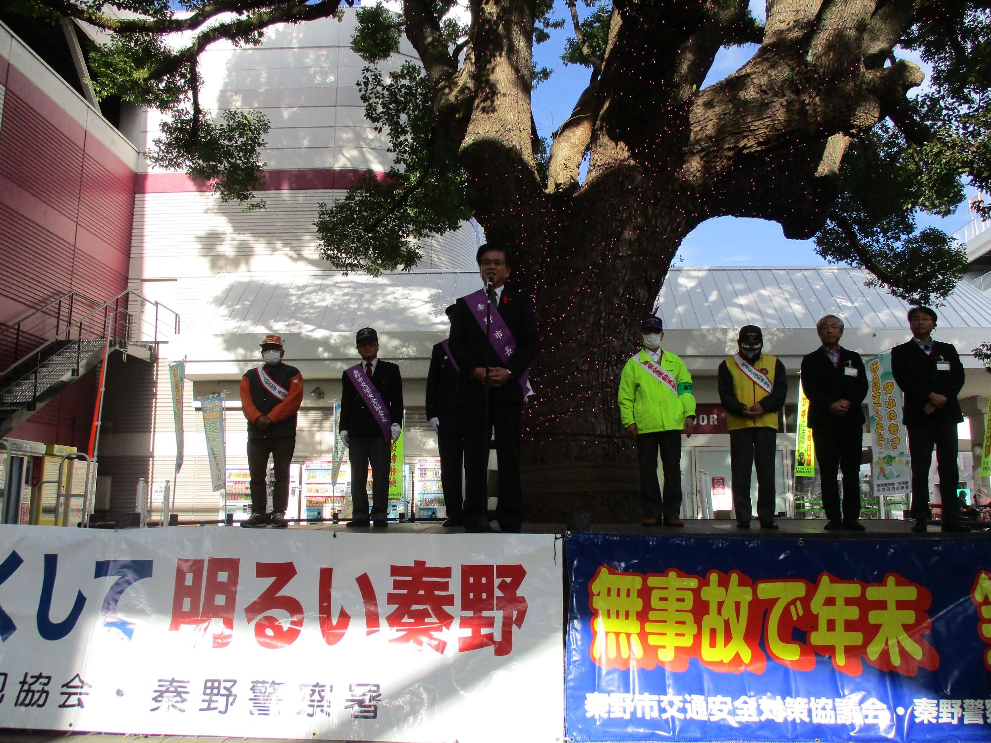 大きな幹の木の前の白と青の横断幕がある広場のステージの上で複数のスーツ姿の男性が並んでおり、中央に立つ市長がマイクを持って挨拶している様子を捉えた写真