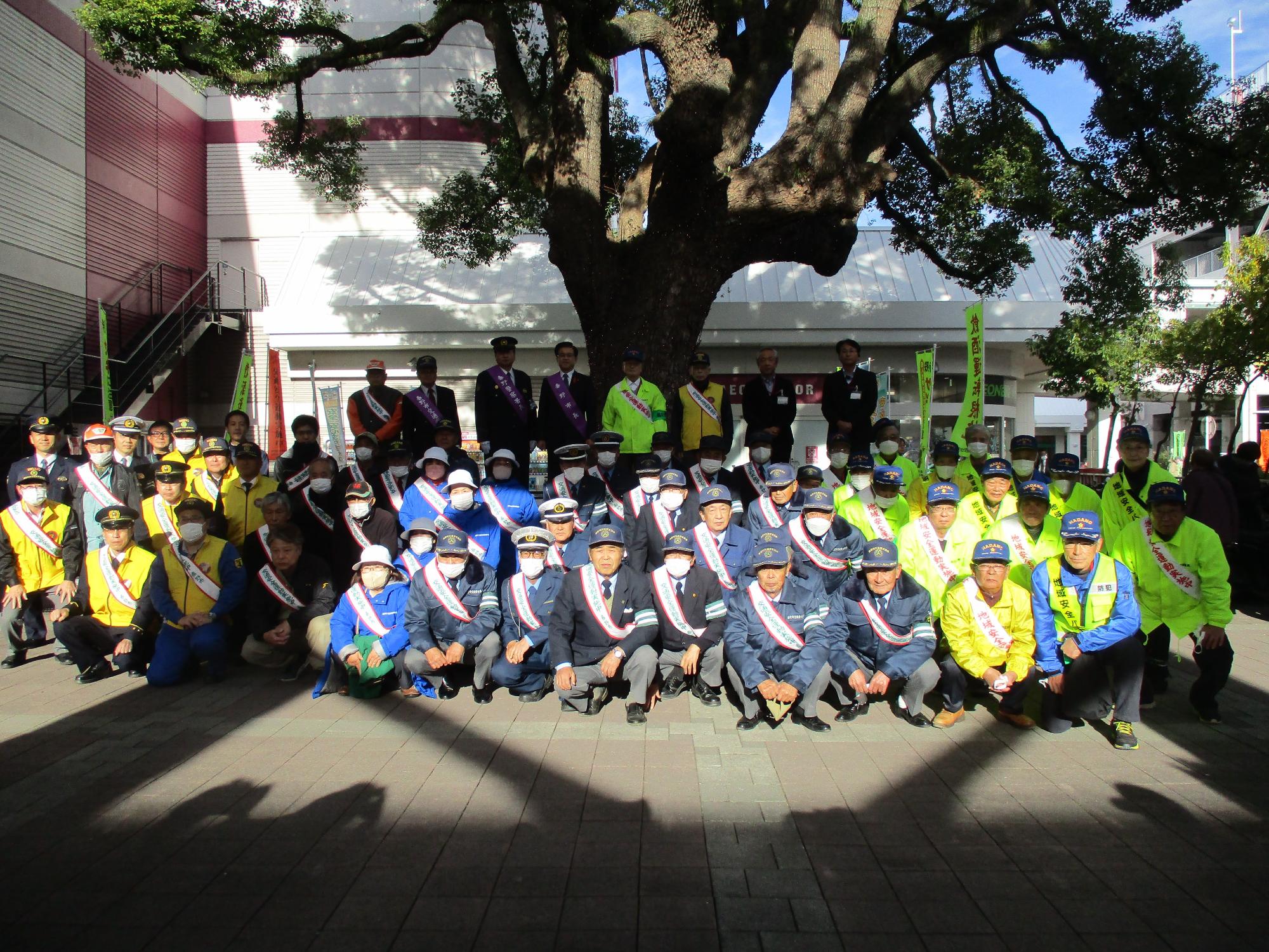 紺色のキャップにジャンパーを着た人達や、黄色や蛍光色のジャンパーを着用した沢山の人々、ステージの上に襷をかけた市長や7人の関係者が並んで集合写真を撮影している屋外でのイベント風景の写真