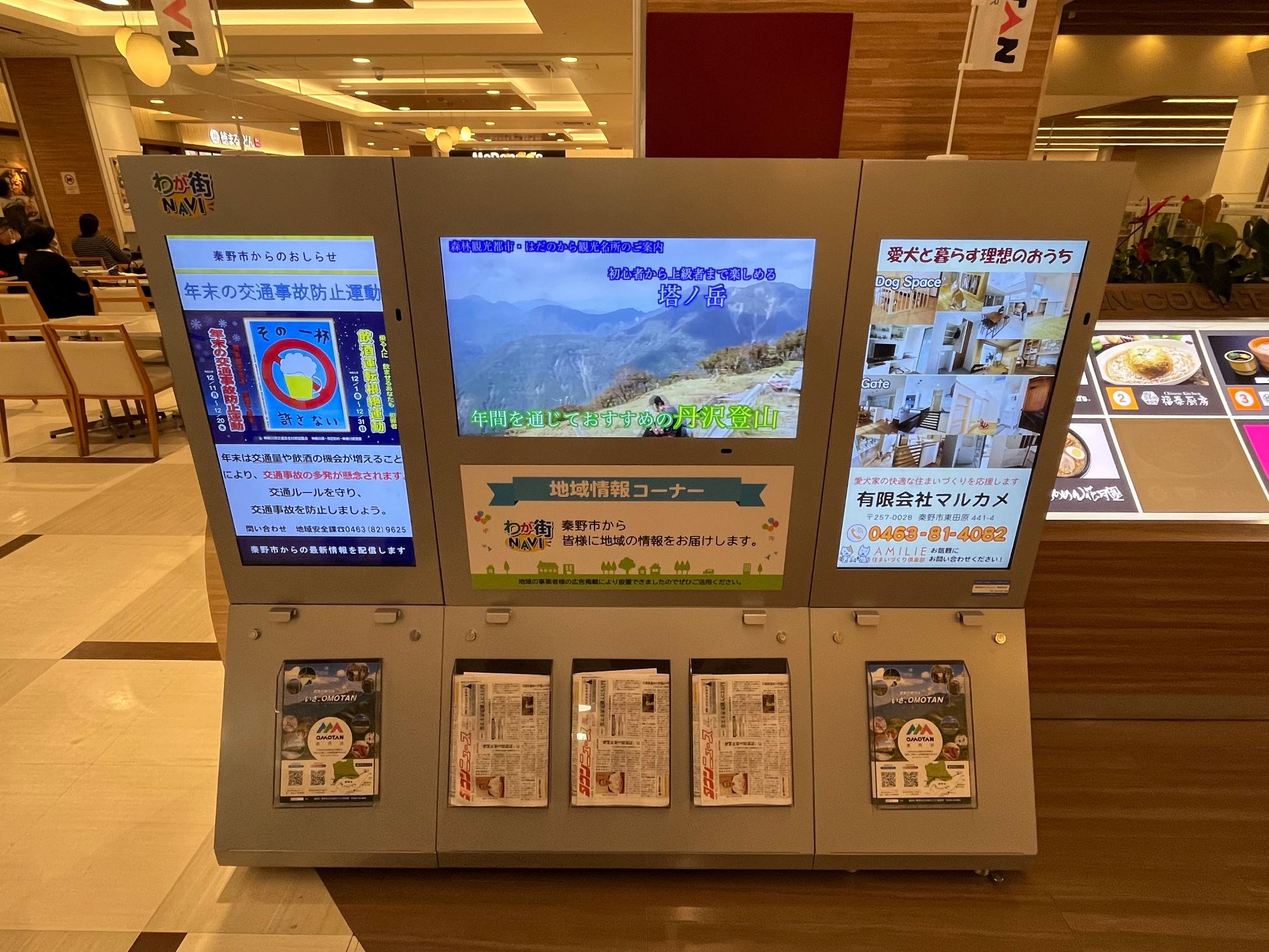 パンフレットが備え付けられている秦野市の地域情報を発信する大型デジタルサイネージで、山や部屋の映像が流れ、左側に「年末の交通事故防止運動」の広告が表示されている写真