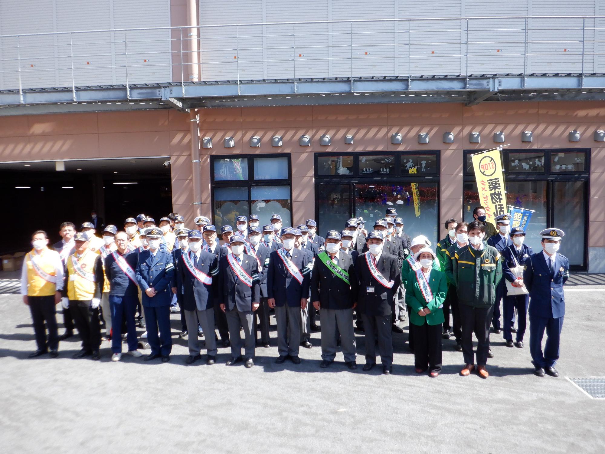大きな建物の前の駐車場で、たすきを掛けたスーツや制服姿の男性たちが、全員マスクを着用して何列にも並び記念撮影をしている様子の写真