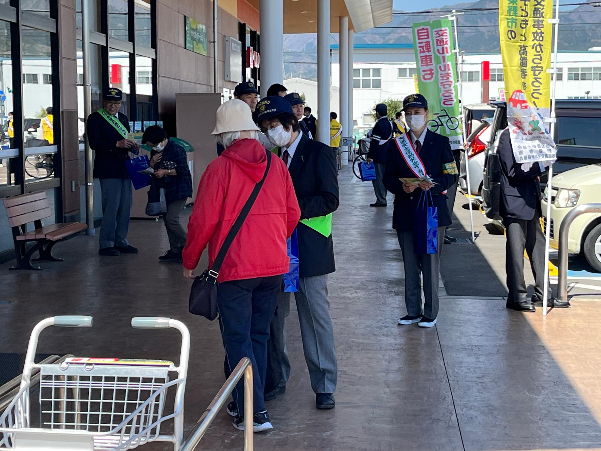 スーパーの店先で、たすきを掛けた制服姿の学生たちが、高齢の買い物客らに交通安全を呼びかける啓発グッズを渡している様子の写真