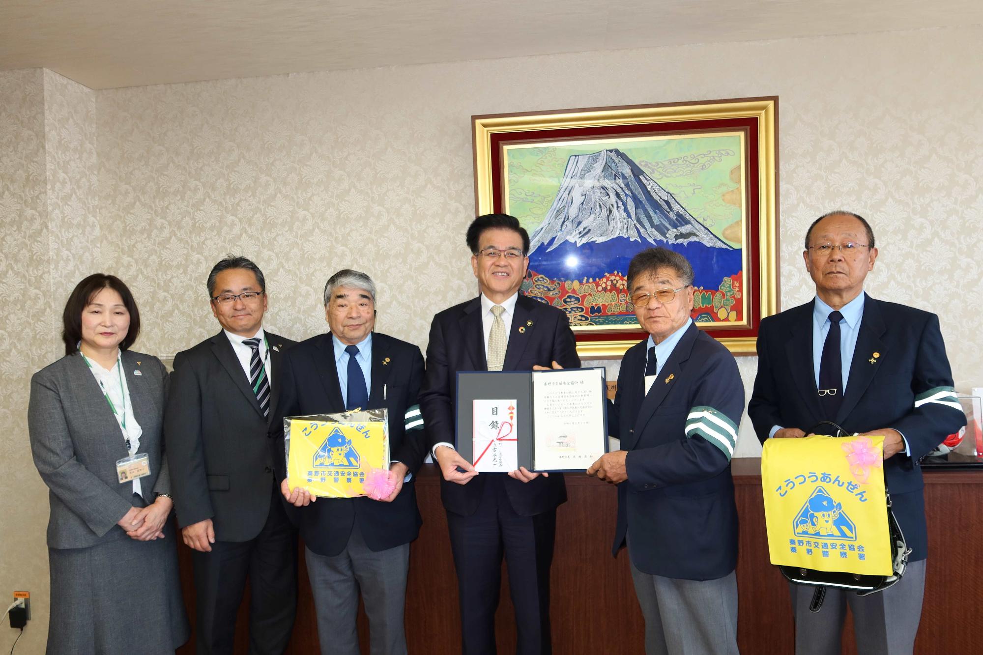 スーツ姿の男女が、交通安全と書かれた黄色いバッグや目録を手に持って記念撮影をしている写真