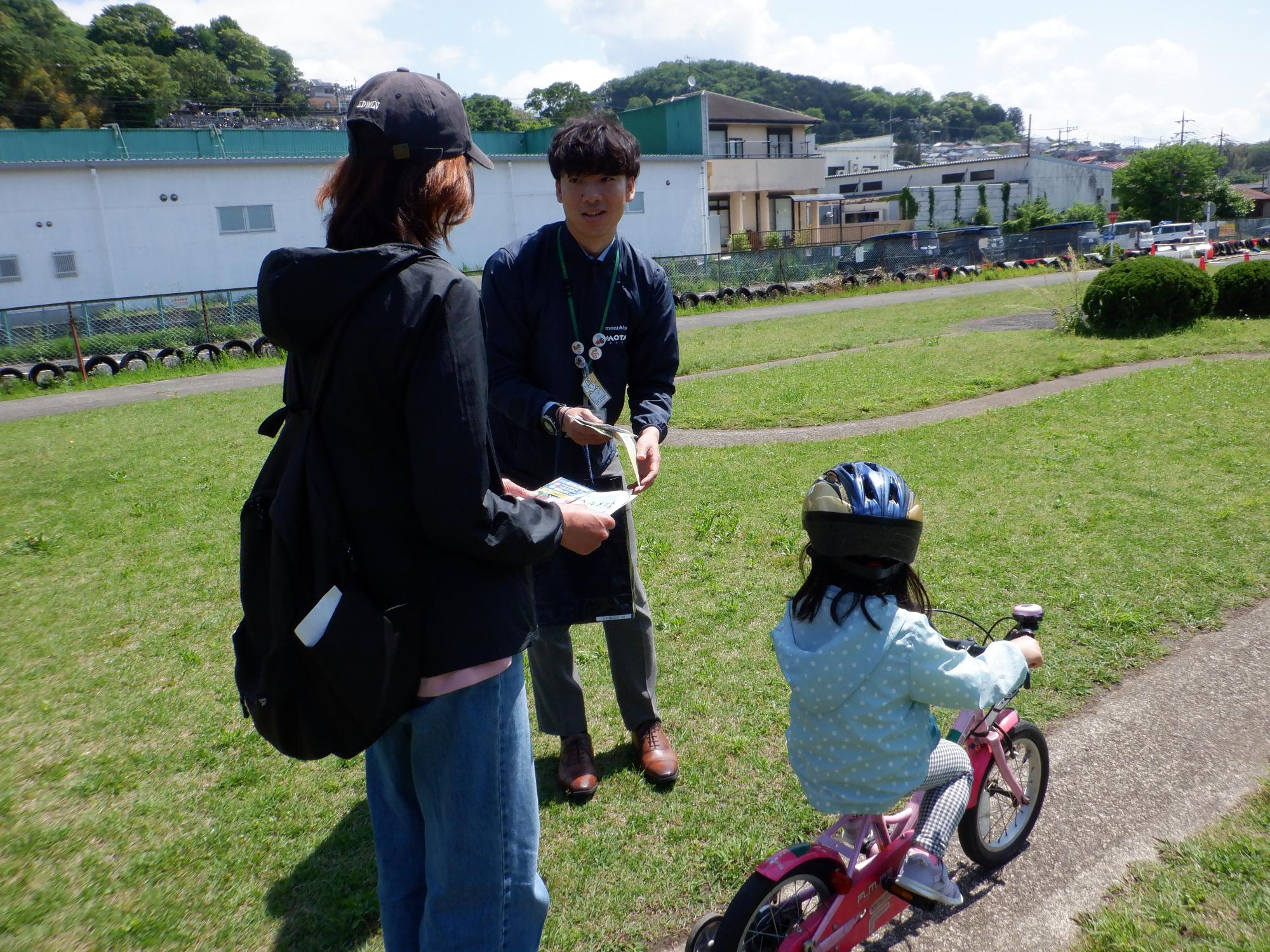 公園で、ヘルメットを被り自転車に乗った子供と親が、男性スタッフから啓発チラシを受け取りながら説明を聞いている様子の写真