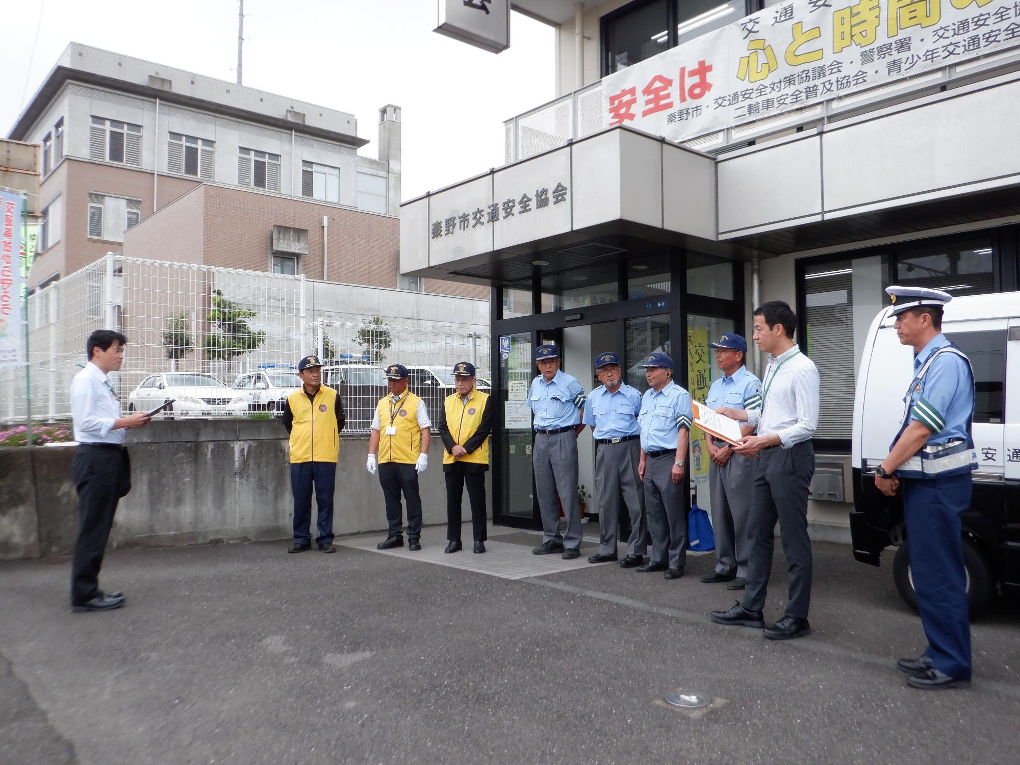秦野市交通安全協会の建物の前で、制服や黄色いベストを着た隊員たちが整列し、出発式のようなセレモニーに臨んでいる様子の写真