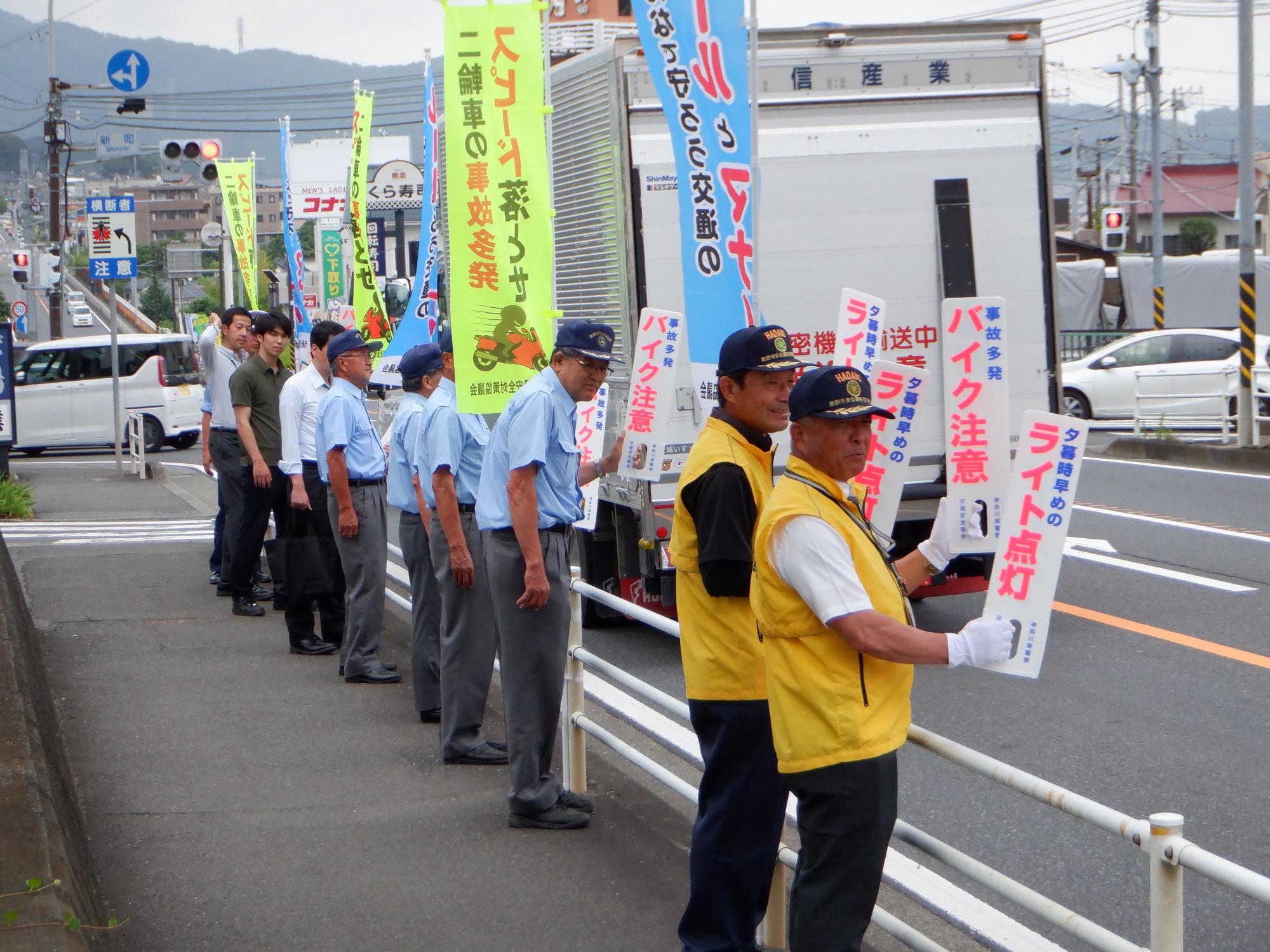 交通量の多い道路沿いに交通安全協会のメンバーらが一列に並び、ドライバーに向けて「バイク注意」などのプラカードを掲げている様子の写真