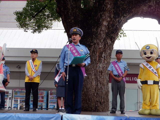 屋外ステージに設置されたマイクの前で、秦野警察署のたすきを掛けた制服姿の警察官が挨拶し、関係者や着ぐるみが見守っている様子の写真