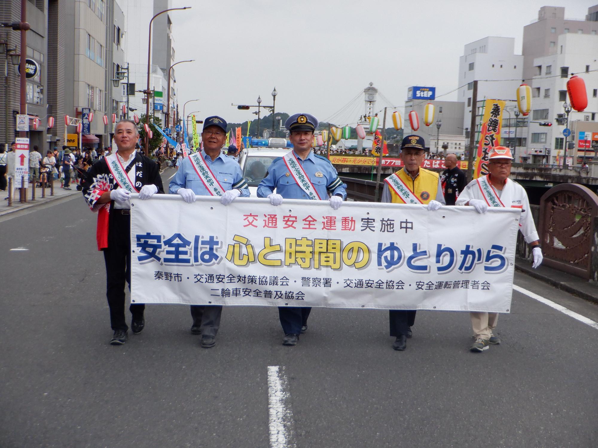 交通安全運動のパレードで、「安全は 心と時間の ゆとりから」と書かれた横断幕を掲げた警察官や関係者らが街中を練り歩いている様子の写真