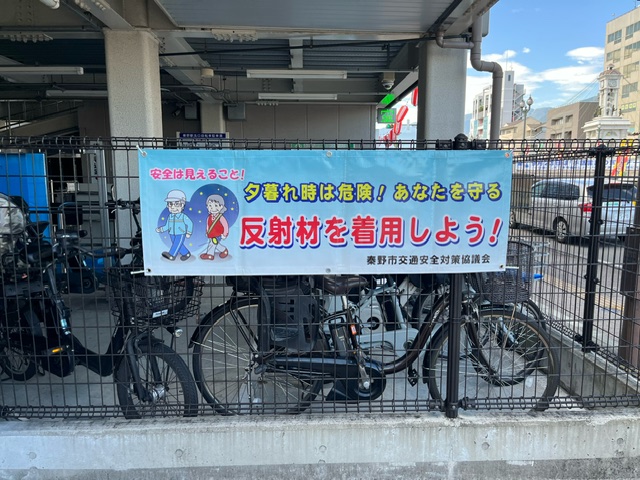 駐輪場のフェンスに、秦野市交通安全対策協議会による「夕暮れ時は危険！反射材を着用しよう！」と書かれた横断幕が掲げられている写真
