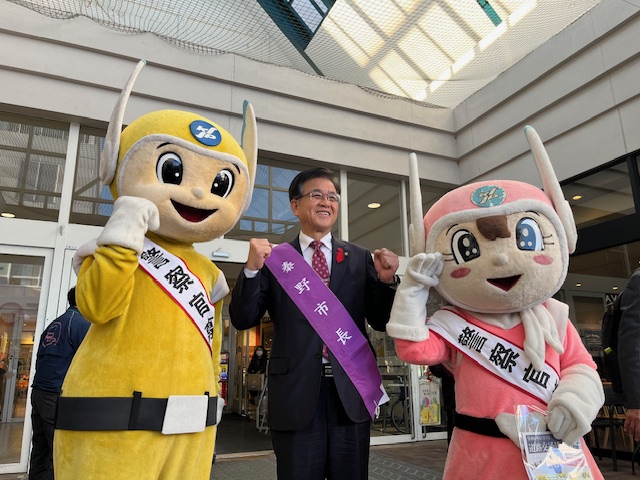 商業施設の入り口で、「秦野市長」と書かれたたすきを掛けた市長が、2体のマスコットキャラクターに挟まれてガッツポーズで記念撮影している写真