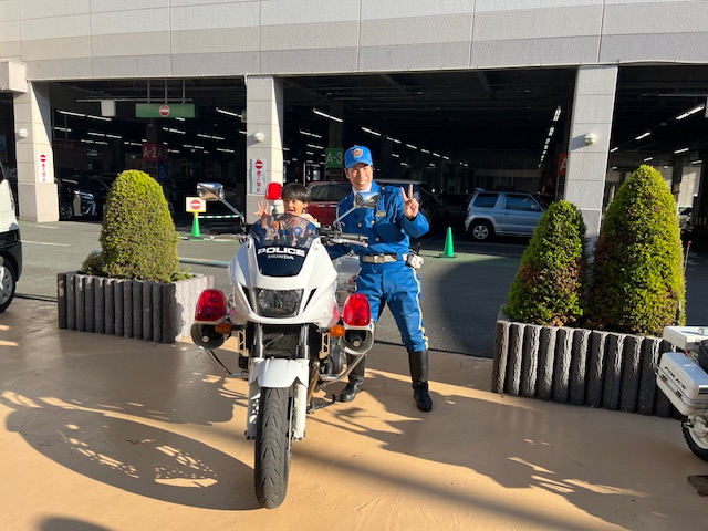 交通安全イベントで、男の子が白バイにまたがり、隣に立つ警察官と一緒に笑顔でピースサインをして記念撮影をしている様子の写真