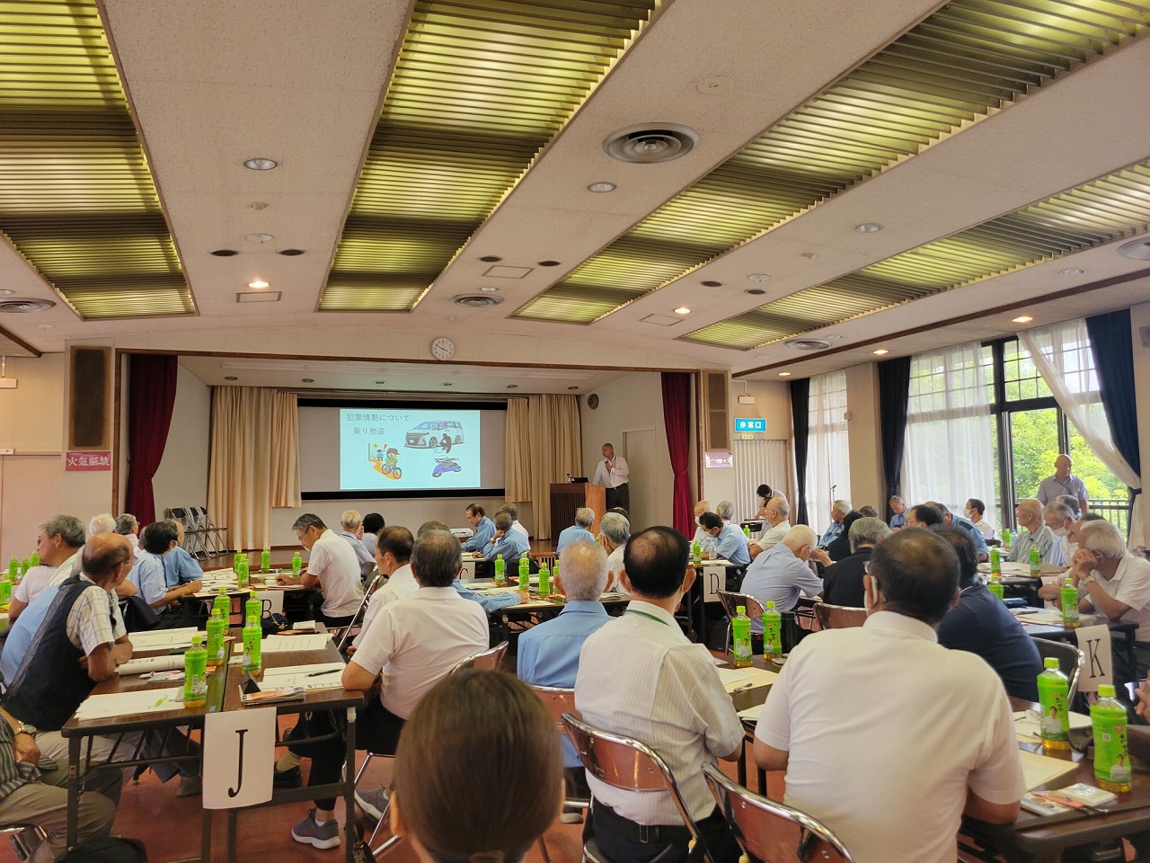 小さなステージのあるホールでの前方に設置されたスクリーンにスライドが映し出され、檀上に立ちマイクを持って話す男性の話を、テーブルごとに分かれて座っている多くの参加者が真剣に聞いている様子の写真