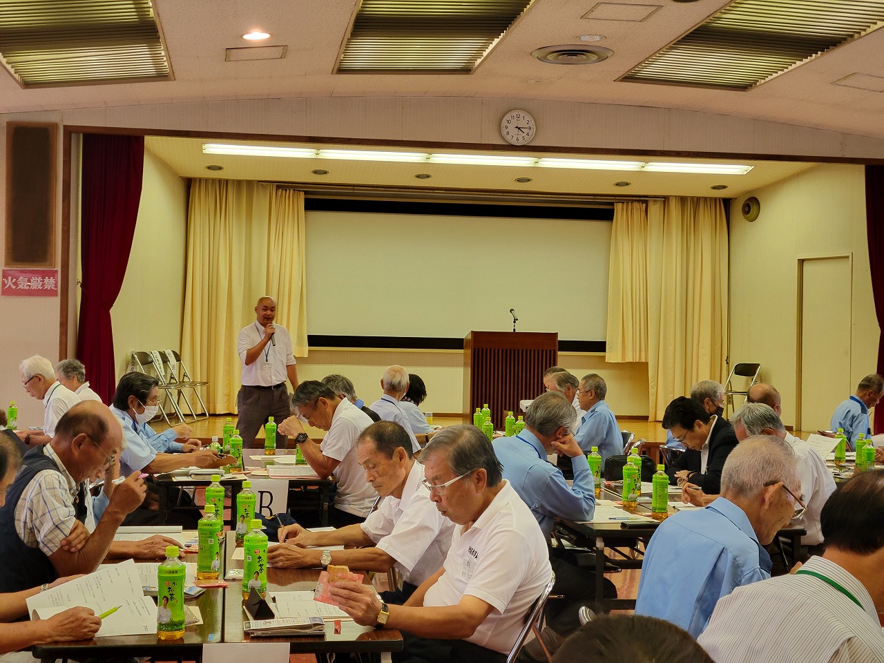 小さなステージのあるホールでお茶のペットボトルが置かれたテーブルごとに多くの参加者が着席しており、ステージ下に立つ男性の話に耳を傾けている会議の様子の写真