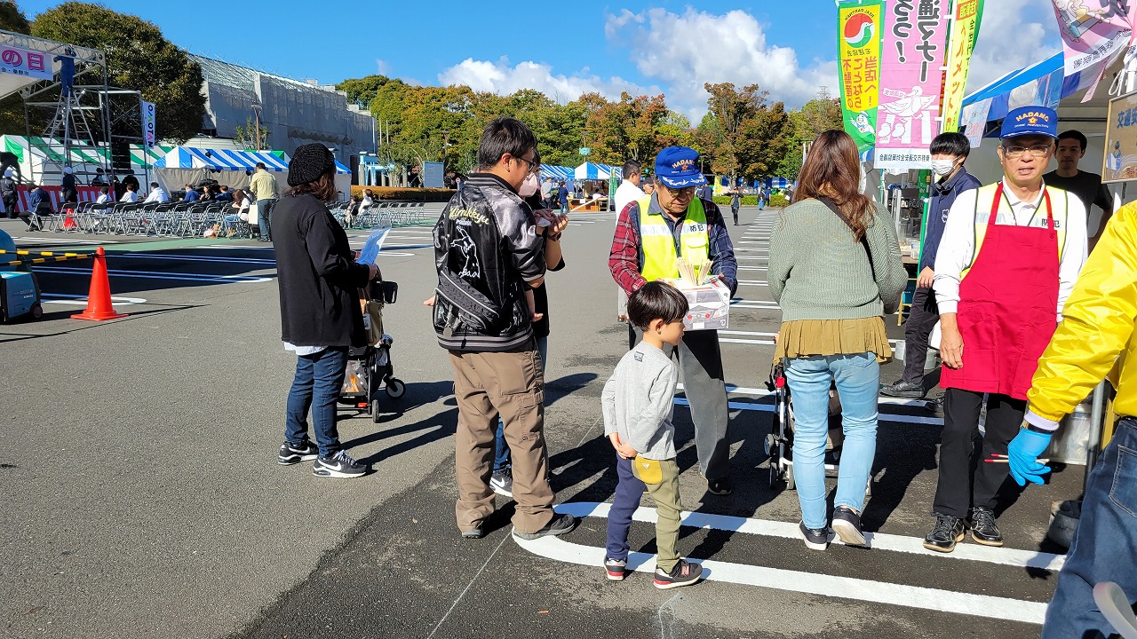 雲が浮かんでいる青い空の駐車場のような広い屋外スペースに青や緑色のストライプのテントが並び、青いキャップを被り黄色のベストを着た男性スタッフが集まっている多くの人々にチラシの様な物を手渡している様子の写真