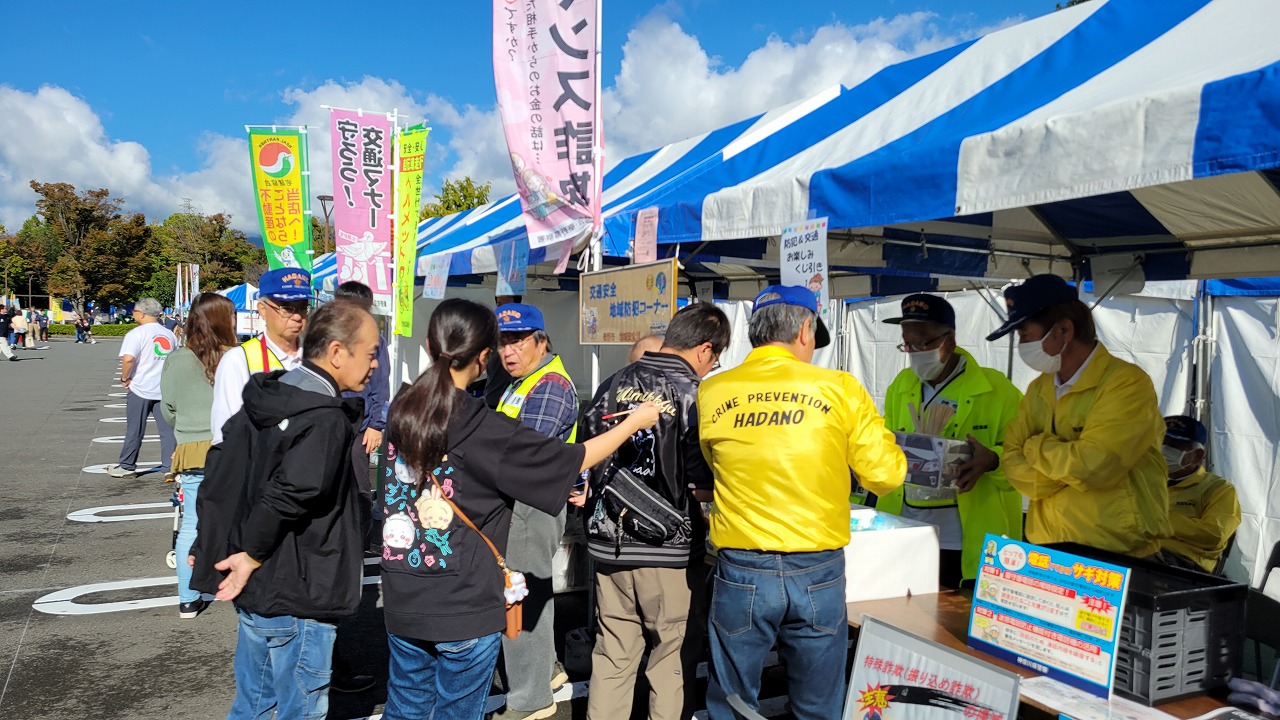 旗がいくつも立てられている青と白の縞模様のテントの前のイベント会場で、青いキャップと黄色いジャンパーを着たスタッフらしき人たちが、訪れた人々と交流している様子の写真