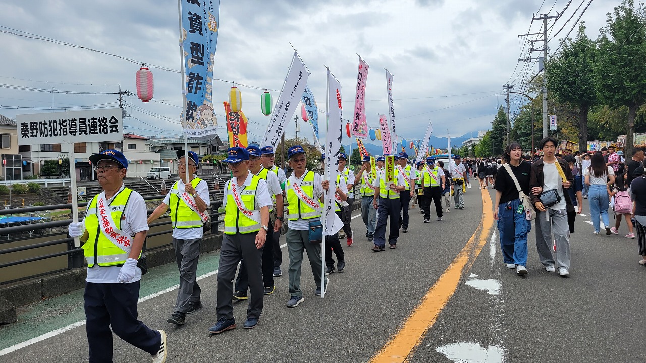 祭りの露店が並んでいる歩行者天国の道路で、タスキをかけ反射ベストを着た人々が旗を掲げながら練り歩いている啓発活動の様子を撮影した写真