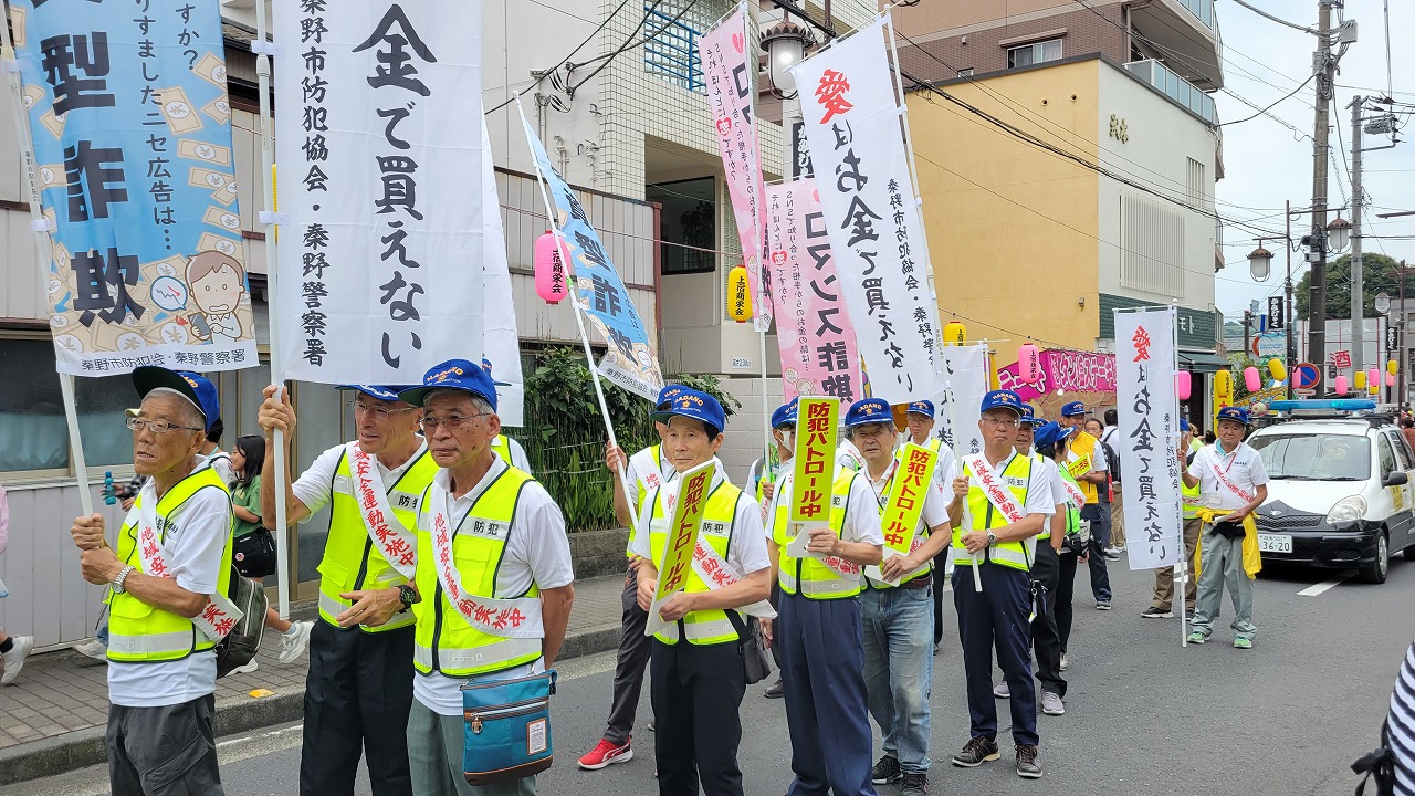 タスキをかけ黄緑色のベストを着た「防犯パトロール中」と書かれた札を持った人々が、のぼり旗を持ちながら市街地の道路をパレードしている防犯啓発活動の様子の写真