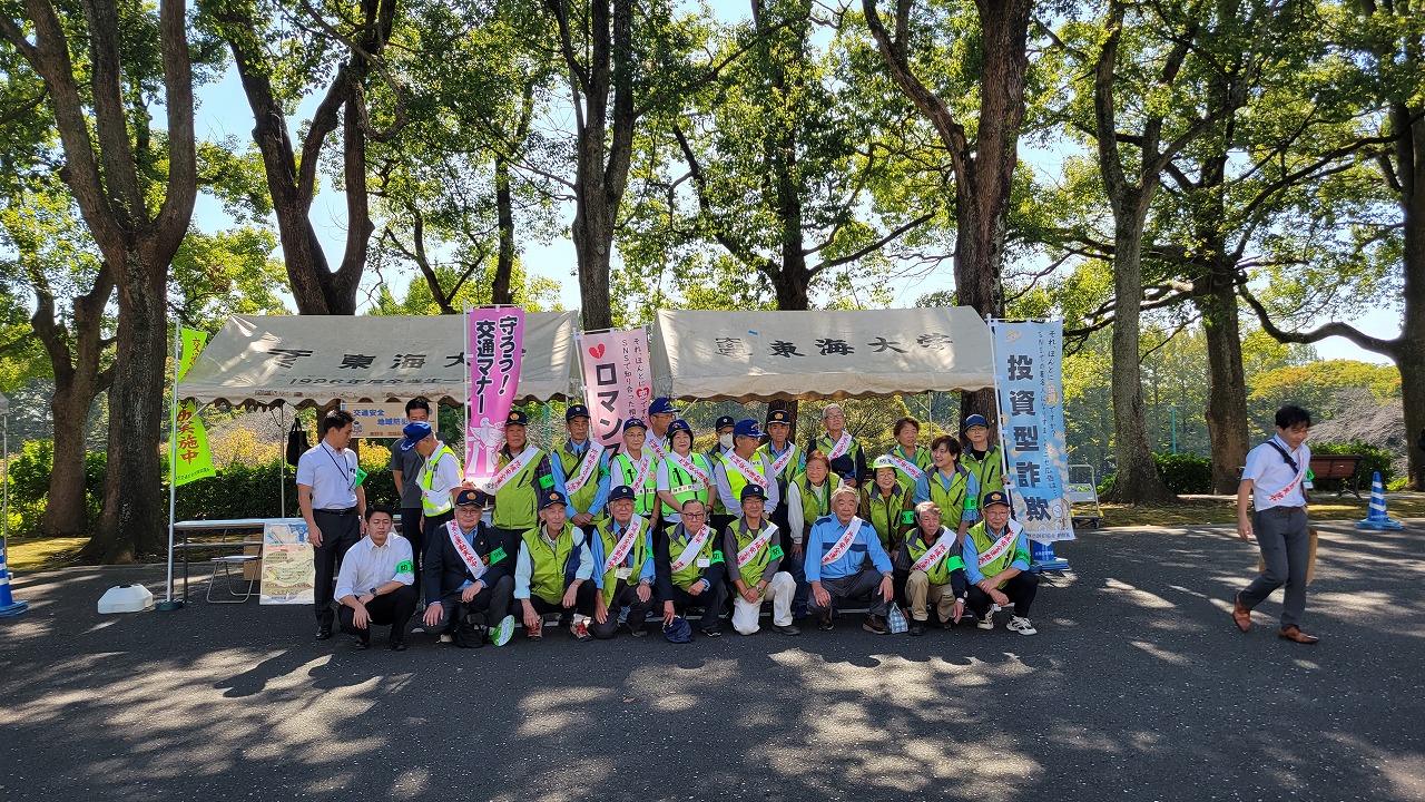 大きな木の下にテントが2台設置された屋外イベント会場で、黄色のベストとタスキをかけた大勢の人々がカメラに向かって並んでいる集合写真